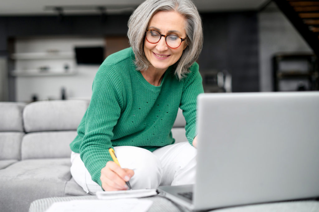 Confident mature woman using laptop computer for remote work, watching webinar and taking notes sitting at home. Contemporary senior female online teacher holding video conference and making marks.